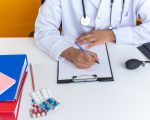 Doctor writing prescription on clipboard at medical desk with pills and stethoscope